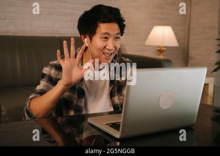 Young Asian male entrepreneur in casual clothing and earphone waving hand while starting video call meeting at late night from home using laptop Stock Photo