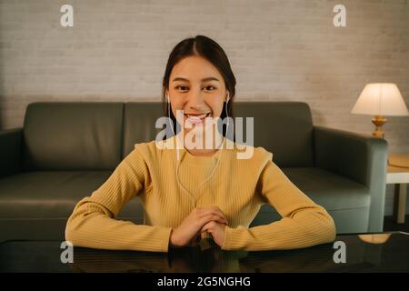 Portrait of smiling young successful Asian woman on video call using headphones sitting focused and concentrated while working late from home at night Stock Photo