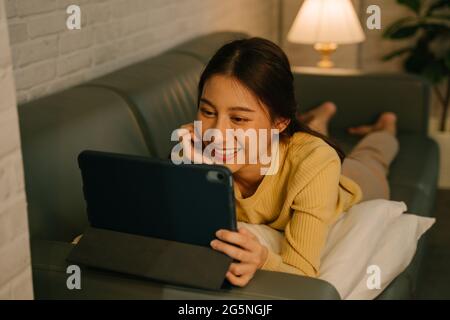Young Asian woman relaxing at home, lying on couch on stomach with pillows using digital tablet and enjoying movies at home during late night Stock Photo