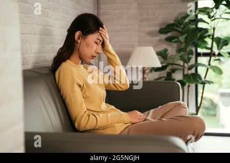 Side view of sad, upset and sick young Asian female in casual clothing sitting with hand on head on couch at home suffering from headache Stock Photo