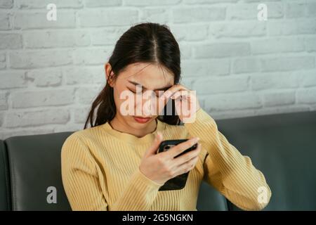 Young Asian woman rubbing eyes due to feeling of discomfort from using smartphone for long hours without rest and break at home Stock Photo