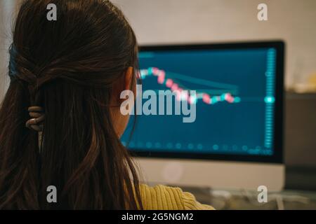 Rear view of young Asian female broker working and reading stock market graphs and data using computer at home with concentration Stock Photo