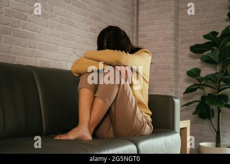 Young woman sitting on couch at home hiding face and hugging knees while feeling depressed and lonely Stock Photo