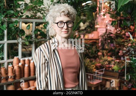 Portrait of young saleswoman in eyeglasses looking at camera while working in the garden with plants Stock Photo