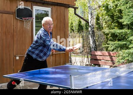 Senior mature grey caucasian old male person making strong ball shot playing ping pong table tennis at home yard garden outdoors on sunny day. Elderly Stock Photo