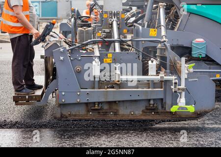 A road worker operates an asphalt paver and pours asphalt concrete while standing behind a control panel on a metal step on a summer day. Stock Photo