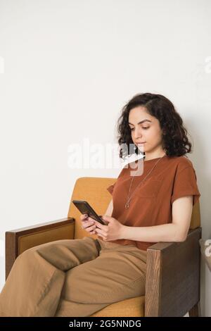 Young woman sitting on armchair and typing a message on her mobile phone Stock Photo