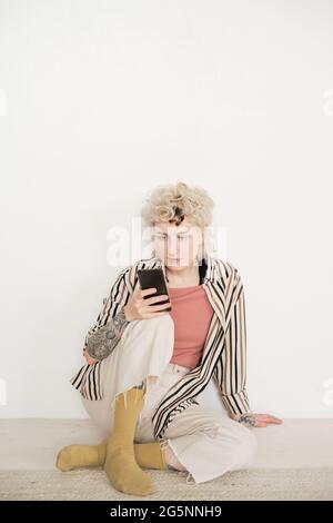 Young woman in stylish clothes sitting on the floor in the room and using her mobile phone Stock Photo
