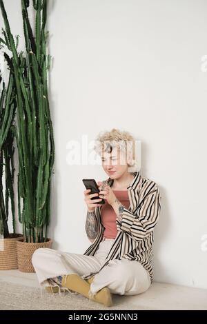Young woman sitting on the floor in the room with plants and reading a book online using her mobile phone Stock Photo