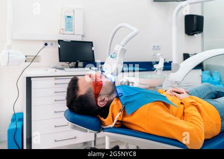 Man lying down in dental chair and wearing protective glasses using tooth whitening machine. Stock Photo