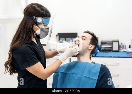 Female dentist wearing face shield isolation mask working with male patient and putting teeth covers. Stock Photo