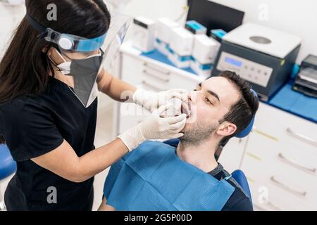 Female dentist wearing face shield isolation mask working with male patient and putting teeth covers. Stock Photo