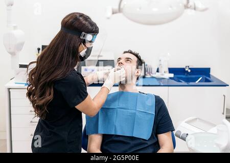 Female dentist wearing face shield isolation mask working with male patient and putting teeth covers. Stock Photo