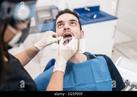 Female dentist wearing face shield isolation mask working with male patient and putting teeth covers. Stock Photo