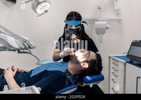 Female dentist wearing protective face mask working with young man during checkup revision. Stock Photo