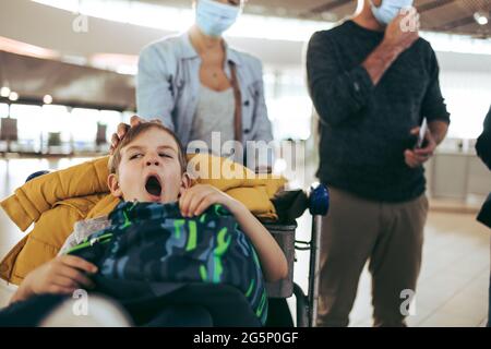 Boy yawning while sitting on luggage trolley at airport with parents in background. Small boy feeling tired while waiting at airport with parents. Stock Photo