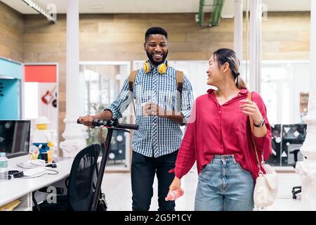 Young asian woman talking with her african coworker in the office. The man is carrying his electric scooter. Stock Photo