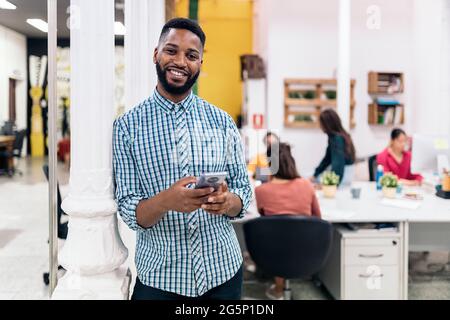 Smiley african office worker holding his mobile phone and looking at camera. Co-working concept. Stock Photo