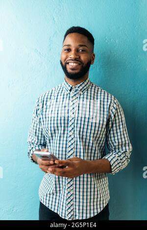 Smiley african office worker holding his mobile phone and looking at camera against blue background. Stock Photo