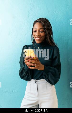 Smiley african office worker holding her mobile phone and typing against blue background. Stock Photo