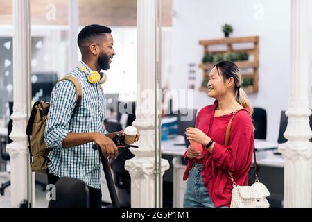 Young asian woman talking with her african coworker in the office. The man is carrying his electric scooter. Stock Photo