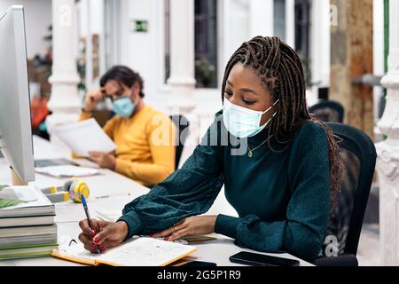 Focused black woman with face mask sitting in her desk and doing paper work next to other coworkers. Office concept. Stock Photo