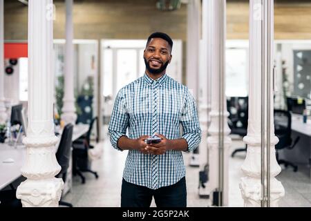 Smiley african office worker holding his mobile phone and looking at camera. Co-working concept. Stock Photo