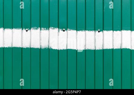 Green metal fence with vertical corrugated stripes, with a horizontal stripe of white paint in the middle and with three bolts Stock Photo