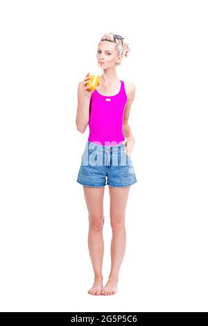 Vertical full length studio portrait of modern young Caucasian woman wearing summer outfit drinking fresh orange juice, white background Stock Photo