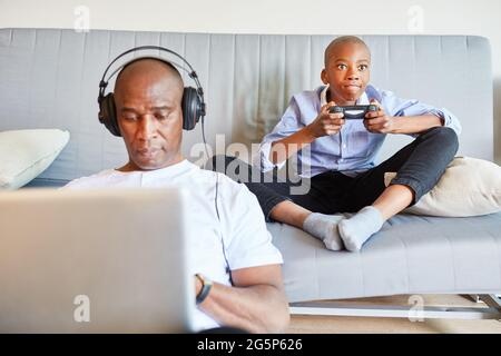 Child on the sofa playing video game and business man using laptop PC video chat in the home office Stock Photo