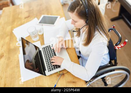 Woman in wheelchair with headset having a video conference on laptop computer in home office Stock Photo