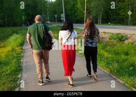 A guy and two girls are walking along the road. Young people are walking down the street. Three people shot from the back. Walk of young people around Stock Photo