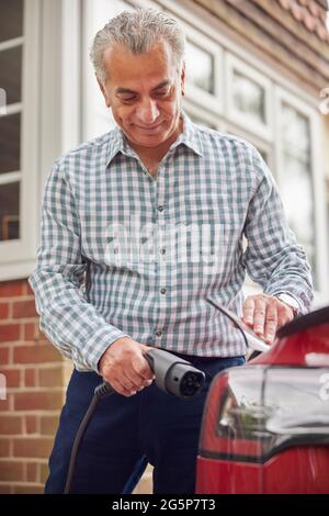 Mature Man Attaching Charging Cable To Environmentally Friendly Zero Emission Electric Car At Home Stock Photo