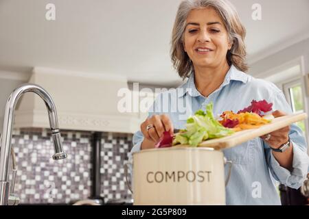 Mature Woman In Kitchen Making Compost Scraping Vegetable Leftovers Into Bin Stock Photo