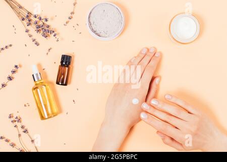 Top view female hands applying cosmetic moisturizer to skin next to natural ingredients for home cosmetics, lavender oil and clay powder on beige back Stock Photo