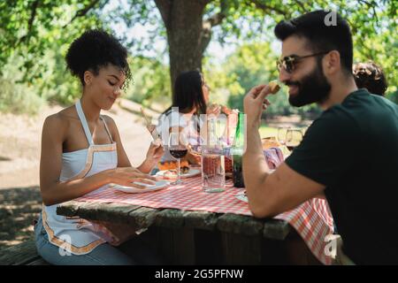 Friends gathering at picnic table in the countryside. Multiracial friends sitting on a wooden table eating skewers of meat and drinking red wine toget Stock Photo