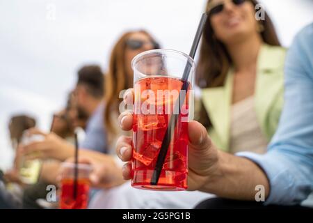 Gathering of best friends having fun talking and drinking cocktails together. Close up of an hand holding an iced spritz drink. Defocused young people Stock Photo
