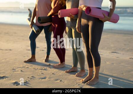 Low section of diverse female friends practicing yoga, at the beach holding yoga mats Stock Photo