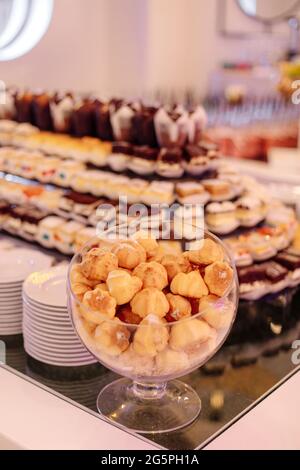 Glass vase with small cakes stands on dessert table, behind it are various cakes and desserts. Wedding event. Service at hotel. Recreation and travel. Stock Photo