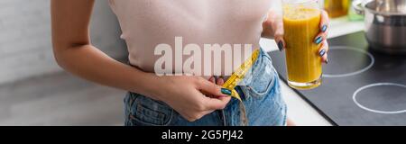 cropped view of young woman holding glass of smoothie while measuring waist, banner Stock Photo