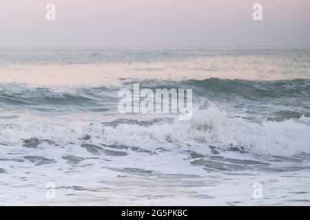 Blue clear sea water and white foam from waves against background of horizon and bright delicate pink dawn sky in the morning. Minimalist seascape. Stock Photo