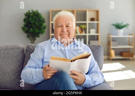 Smiling elderly man or grandfather in blue shirt sitting on sofa and reading book at home Stock Photo