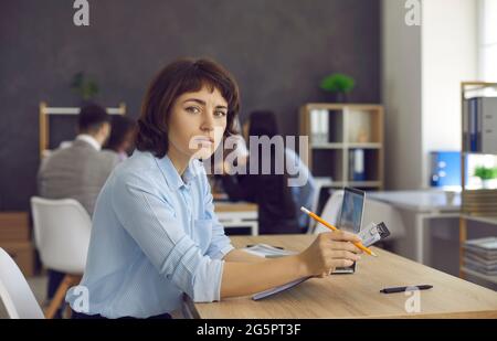 Portrait of businesswoman sitting at office desk, holding clipboard and looking at camera Stock Photo