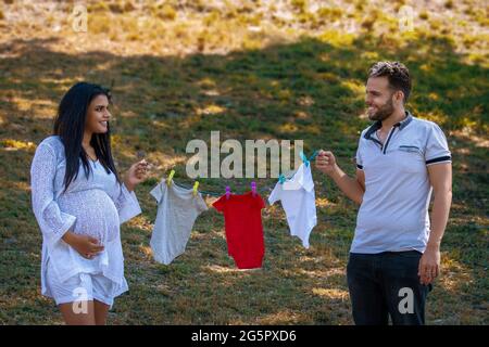 Young couple holding a twine with hanging baby clothes. The young woman is pregnant Stock Photo