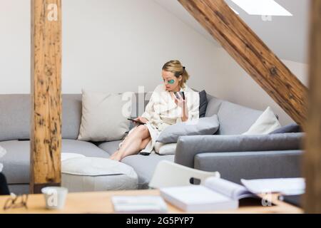 Young female entrepreneur wearing beauty face mask and cosy warm bathrobe in the morning, working remotly from her living room sofa. Remote work from Stock Photo