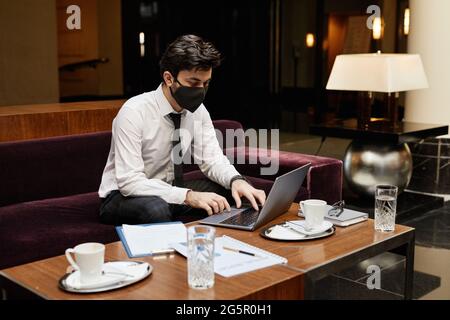 Portrait of young businessman wearing mask while working in hotel lobby, copy space Stock Photo