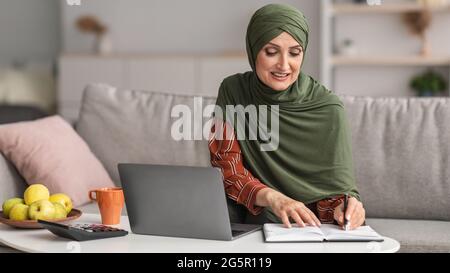 Happy Mature Islamic Lady Using Laptop Taking Notes At Home Stock Photo