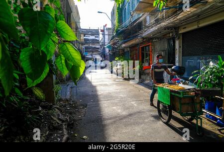 A male food vendor pushes his cart along a sunlit road in Bangkok, Thailand Stock Photo