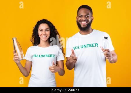 Two Volunteers Holding Glass And Plastic Bottles, Yellow Background Stock Photo