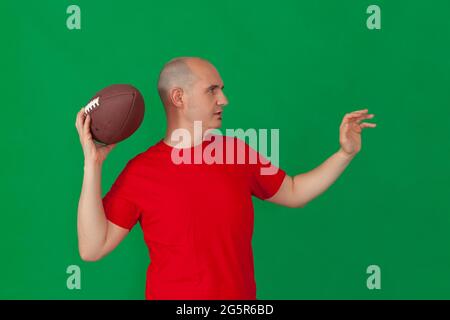 A bald caucasian man dressed in a red t-shirt holding an american football ball positioned as if to throw a forward pass. The background is green. Stock Photo